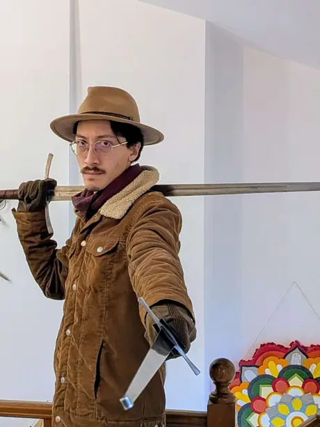 Henrique in a wide-brimmed hat and corduroy jacket, a longsword resting across one shoulder and a dagger held out in the other hand