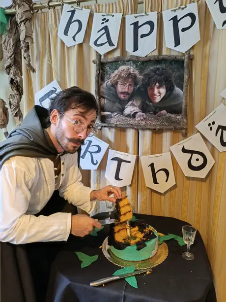Pedro in hobbit costume, cutting a birthday cake iced like the Shire, under a banner lettered in Tengwar