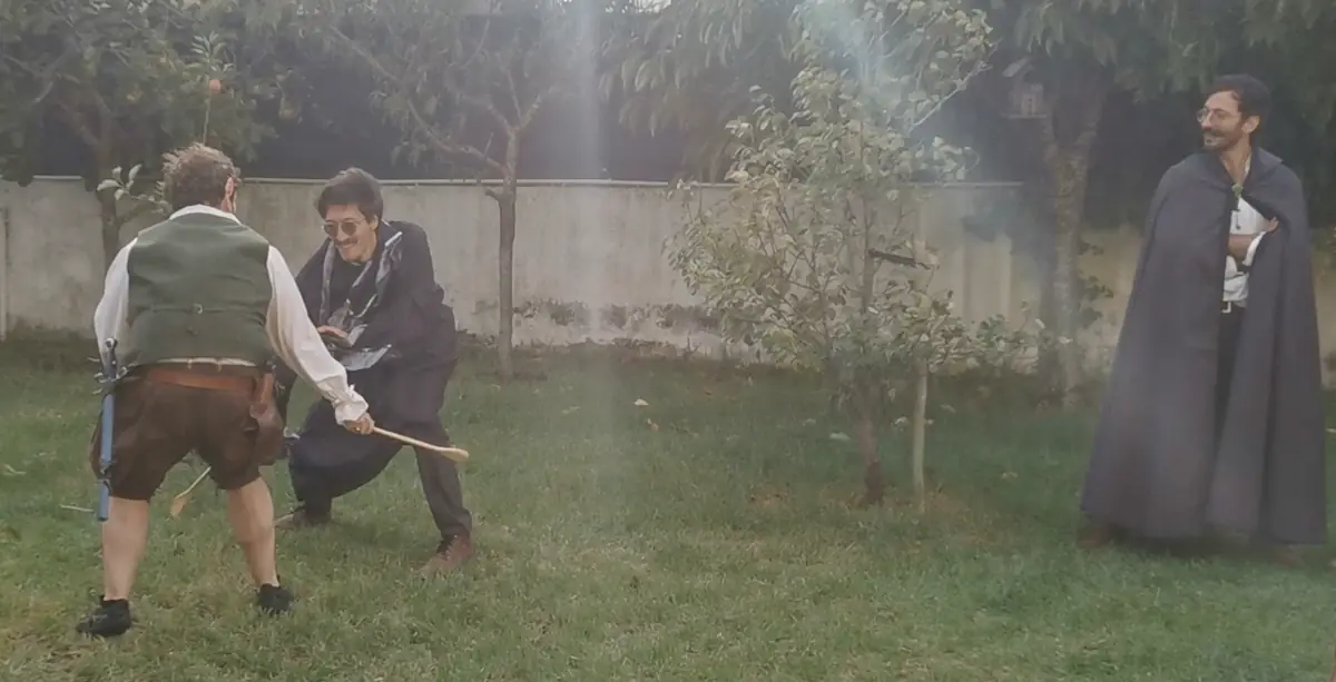 Two of us duelling on a garden lawn in costume, one lunging with a wooden spoon, while a third watches from the side in a grey cape